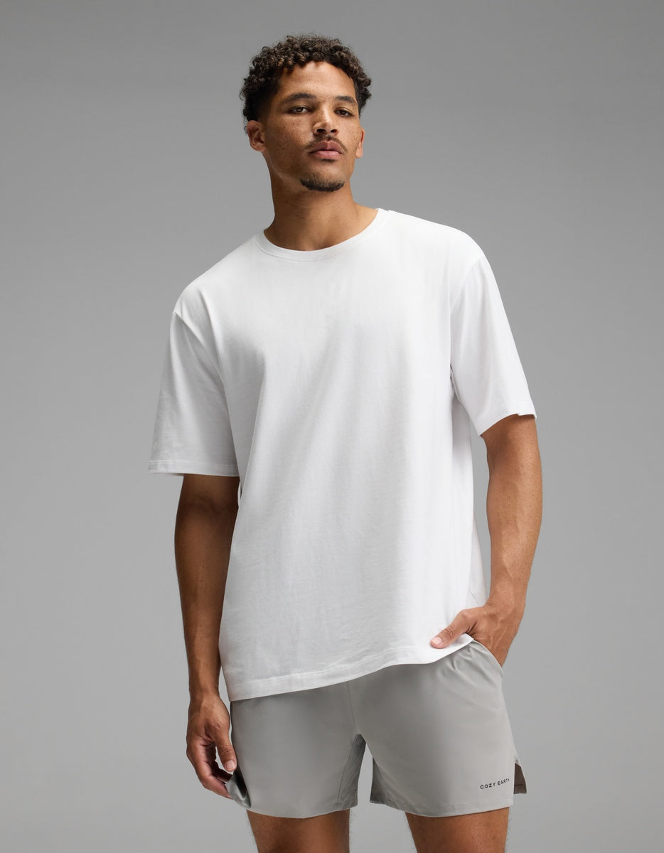 A man stands against a gray background wearing the Cozy Earth Men’s All Day Tee in white and light gray shorts, with one hand in his pocket and a neutral expression.|Color:White