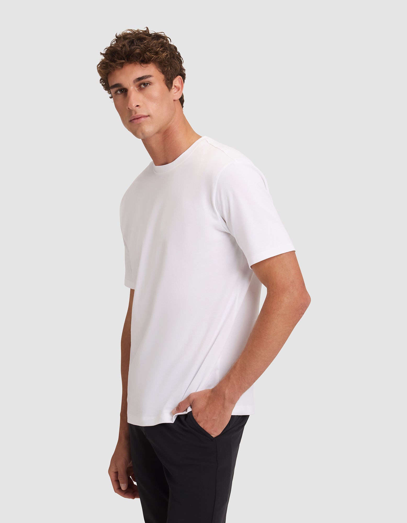 A young man with short curly hair wears the Cozy Earth Men's All Day Tee in plain white and black pants, standing against a light gray background, facing slightly to the side with one hand in his pocket. 
