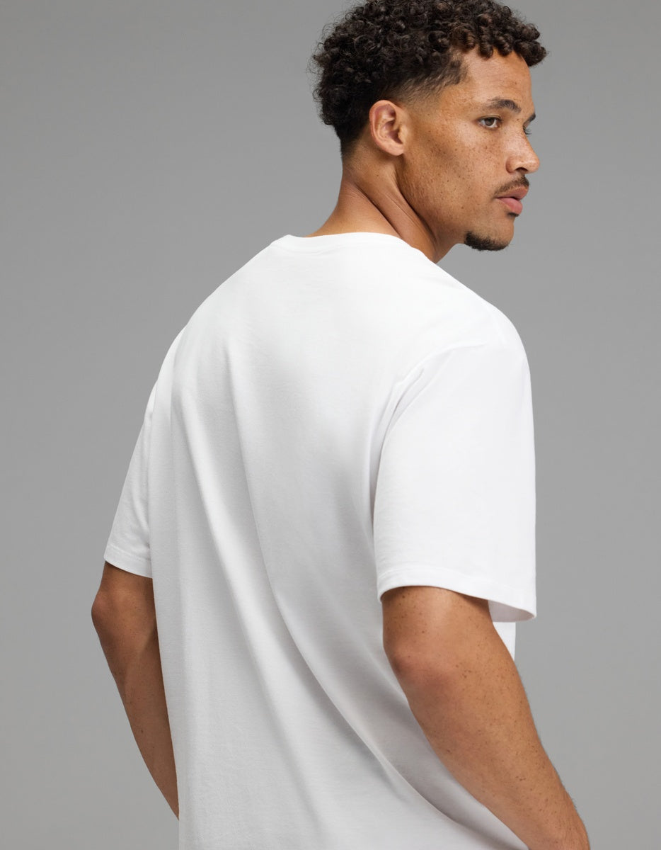 A man with short curly hair and a trimmed beard, wearing the Cozy Earth Men’s All Day Tee in plain white, turns to look over his right shoulder against a neutral gray background.