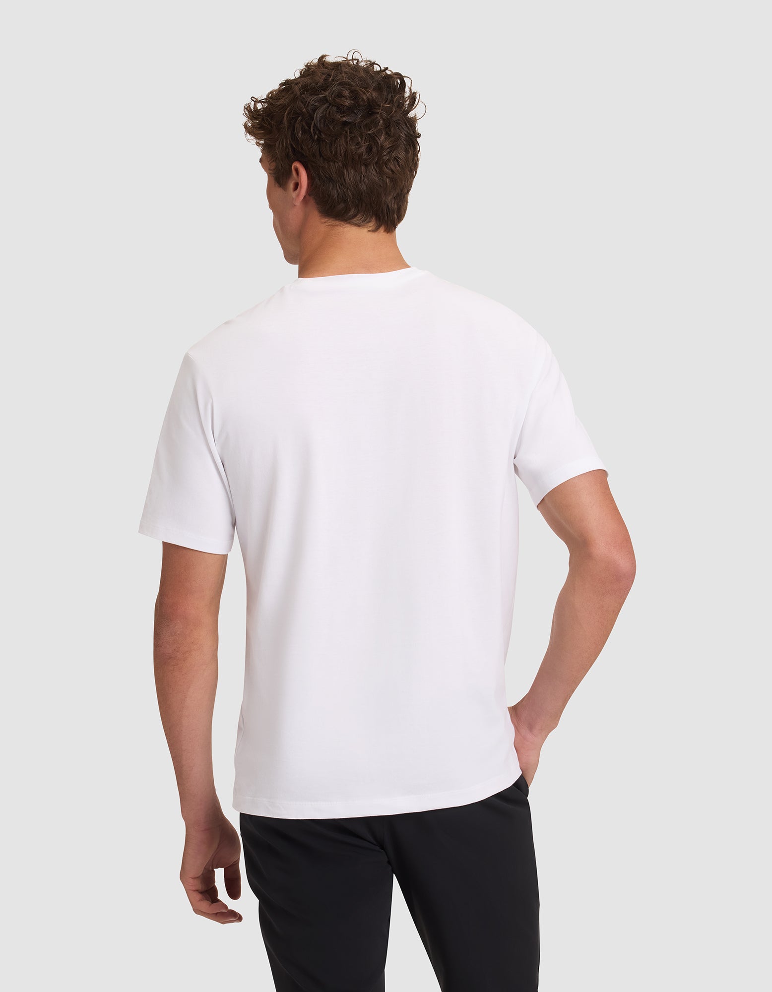 A person with short curly hair is shown from the back, wearing a Cozy Earth Men's All Day Tee in plain white and black pants, standing against a light gray background. 