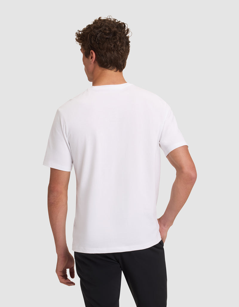 A person with short curly hair is shown from the back, wearing a Cozy Earth Men's All Day Tee in plain white and black pants, standing against a light gray background.