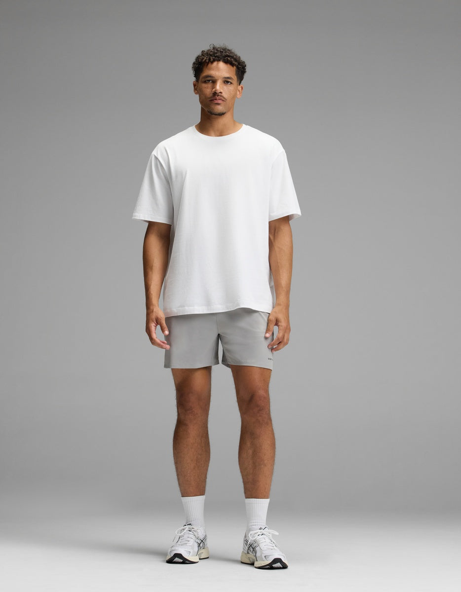 A man stands facing forward against a plain gray background, wearing the Men’s All Day Tee by Cozy Earth, paired with light gray shorts, white socks, and white sneakers.