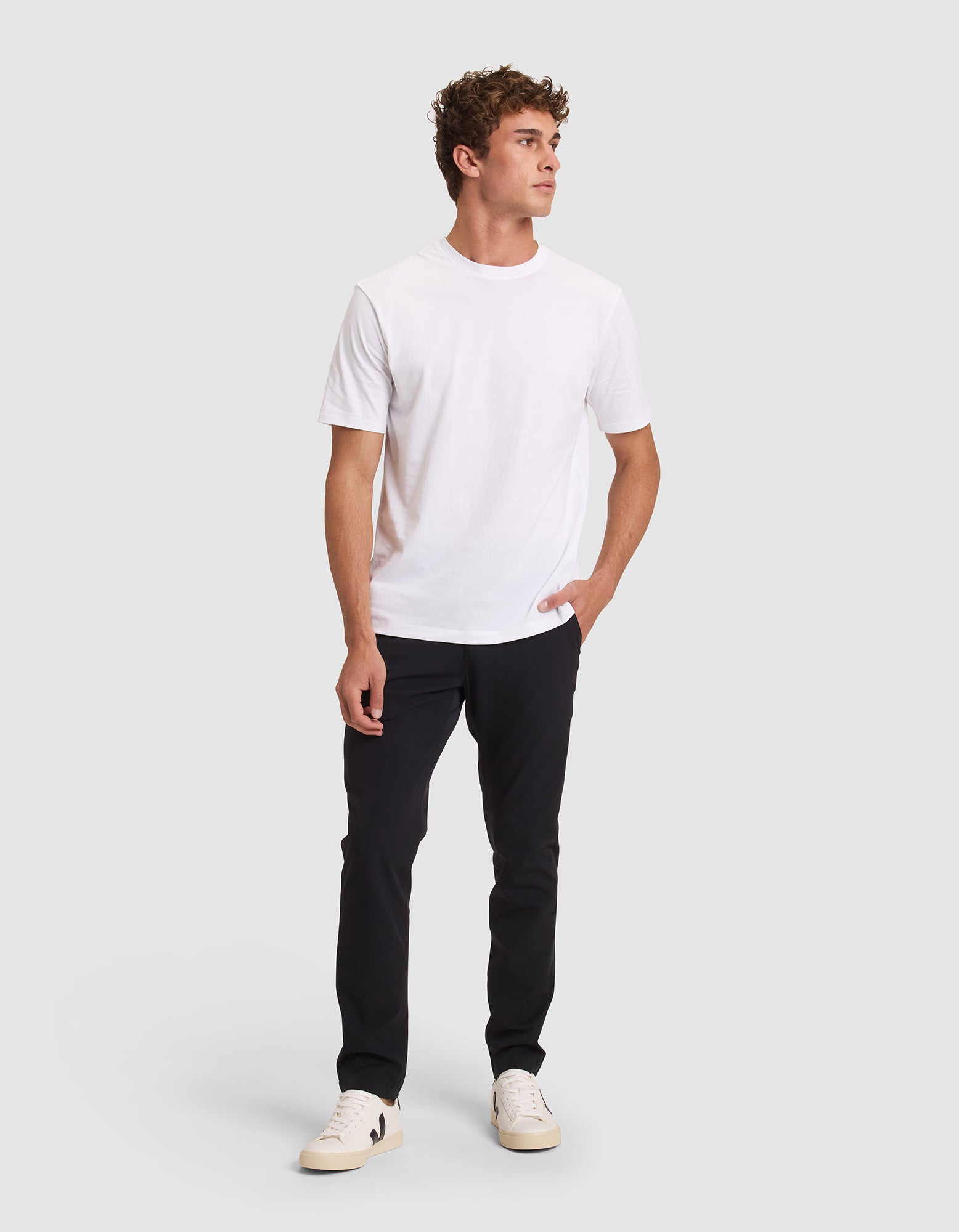 A young man with curly hair models the Cozy Earth Men's All Day Tee—a plain white t-shirt—paired with black pants and white sneakers, standing confidently against a light gray background with one hand in his pocket. 