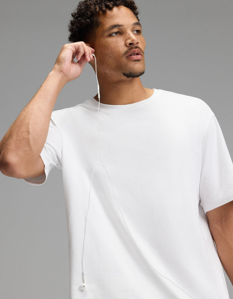 A man wearing the Cozy Earth Men’s All Day Tee in plain white holds an earbud to his ear, gazing off to the side with a neutral expression against a simple gray background.