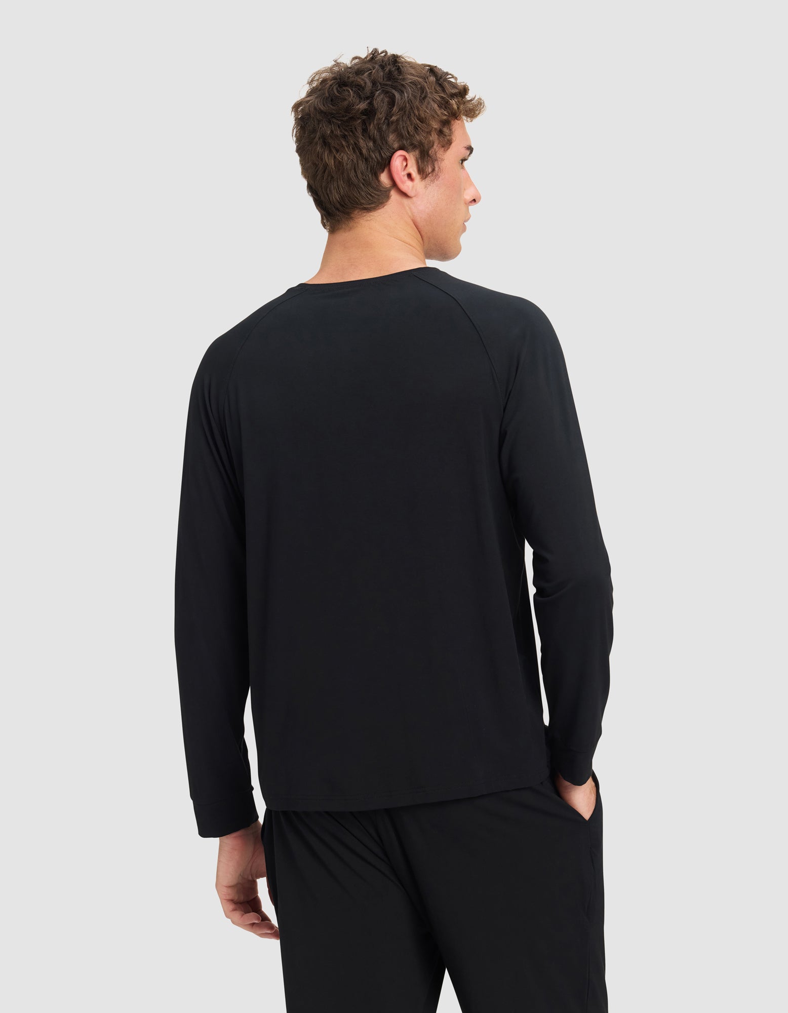 A man with curly brown hair is shown from the back wearing a black Cozy Earth Men's Bamboo Stretch-Knit Long Sleeve shirt and black pants, his left hand in his pocket, against a plain light gray background.