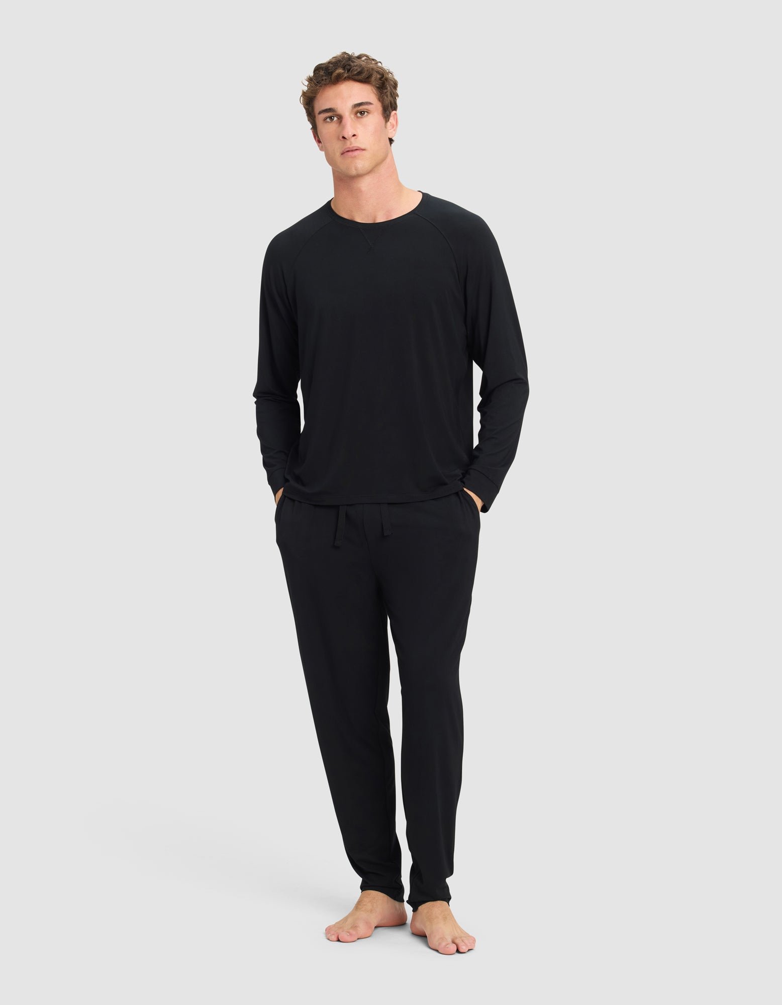 A man stands barefoot against a plain light gray background, wearing the Cozy Earth Men's Bamboo Stretch-Knit Long Sleeve and matching drawstring pants, with his hands in his pockets.|Color:Black
