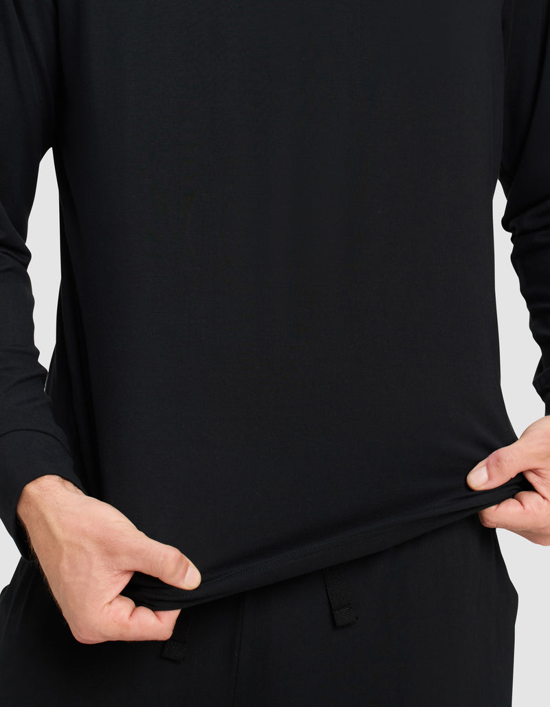 A person wearing the Cozy Earth Men's Bamboo Stretch-Knit Long Sleeve and black pants stretches the shirt's bottom outward with both hands, demonstrating the flexibility of its fabric.