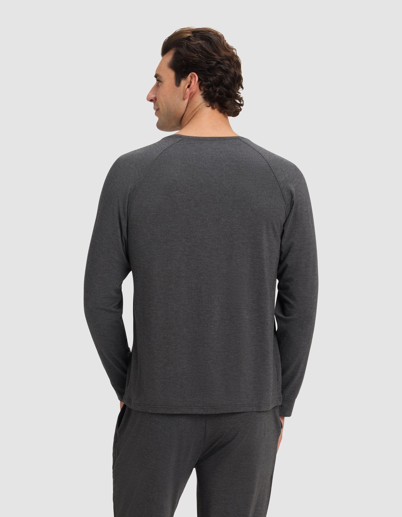 A man with dark hair stands facing away from the camera, wearing the Cozy Earth Men's Bamboo Stretch-Knit Long Sleeve in dark gray, paired with matching pants, set against a plain light background.