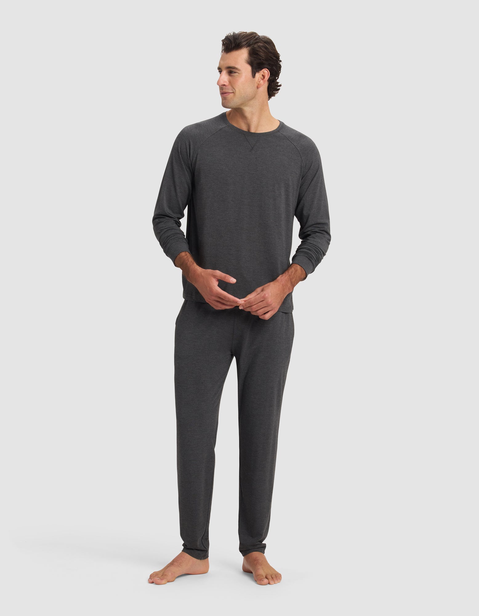 A man stands barefoot against a plain background, wearing Cozy Earth's Men's Bamboo Stretch-Knit Long Sleeve pajama set in dark gray with relaxed fit pants and a crew neck top. He looks to the side, hands clasped loosely.|Color:Charcoal