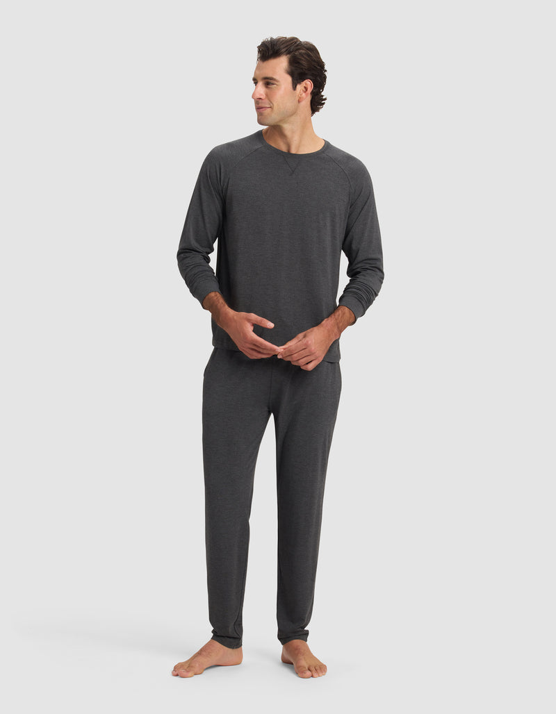 A man stands barefoot against a plain background, wearing Cozy Earth's Men's Bamboo Stretch-Knit Long Sleeve pajama set in dark gray with relaxed fit pants and a crew neck top. He looks to the side, hands clasped loosely.