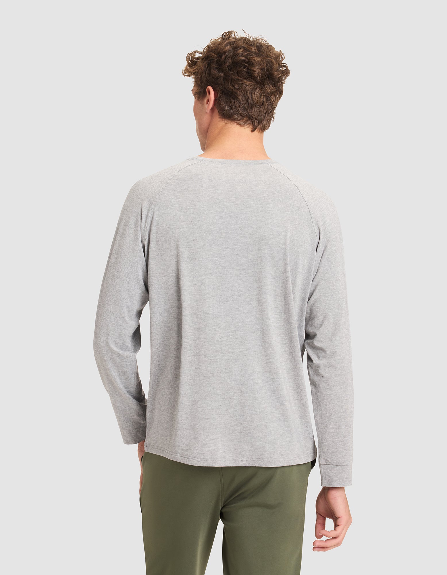 A man with curly hair stands with his back to the camera, wearing a Cozy Earth Men's Bamboo Stretch-Knit Long Sleeve in light gray and olive green pants against a plain light background. 