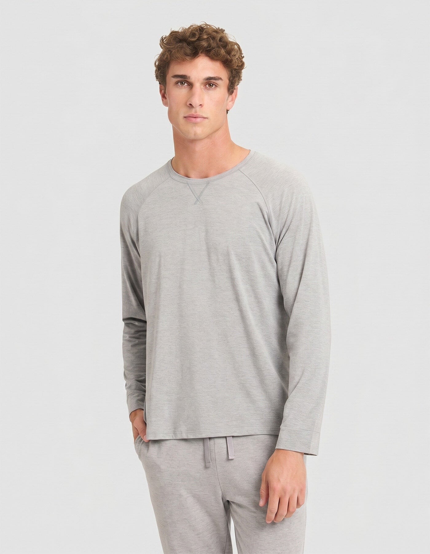 A young man with curly brown hair models the Cozy Earth Men's Bamboo Stretch-Knit Long Sleeve Pajama Set in light gray, standing with one hand in his pocket against a plain, light background.