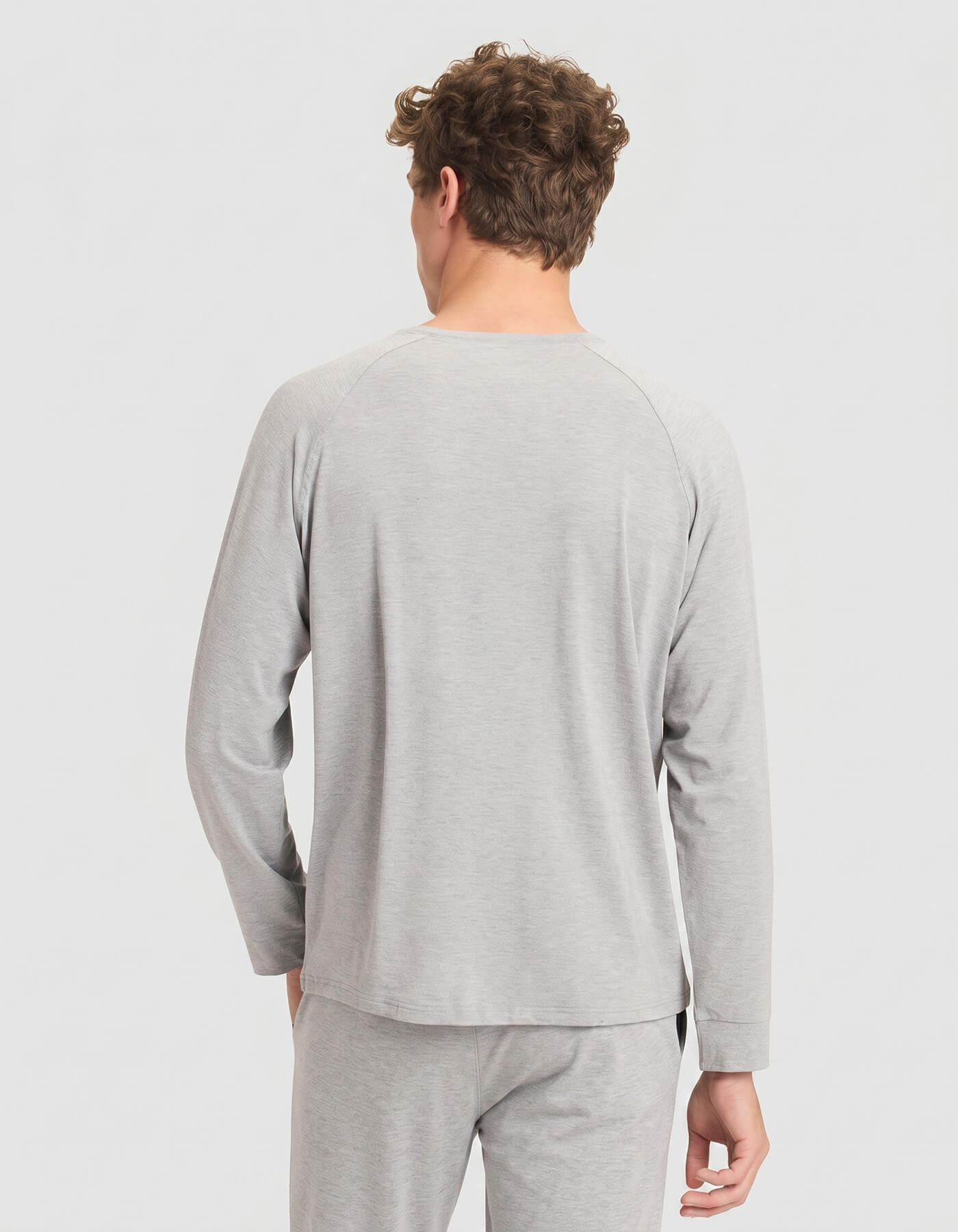 A man with short curly hair is seen from behind wearing the Cozy Earth Men's Bamboo Stretch-Knit Long Sleeve Pajama Set in light gray, standing against a plain light background.