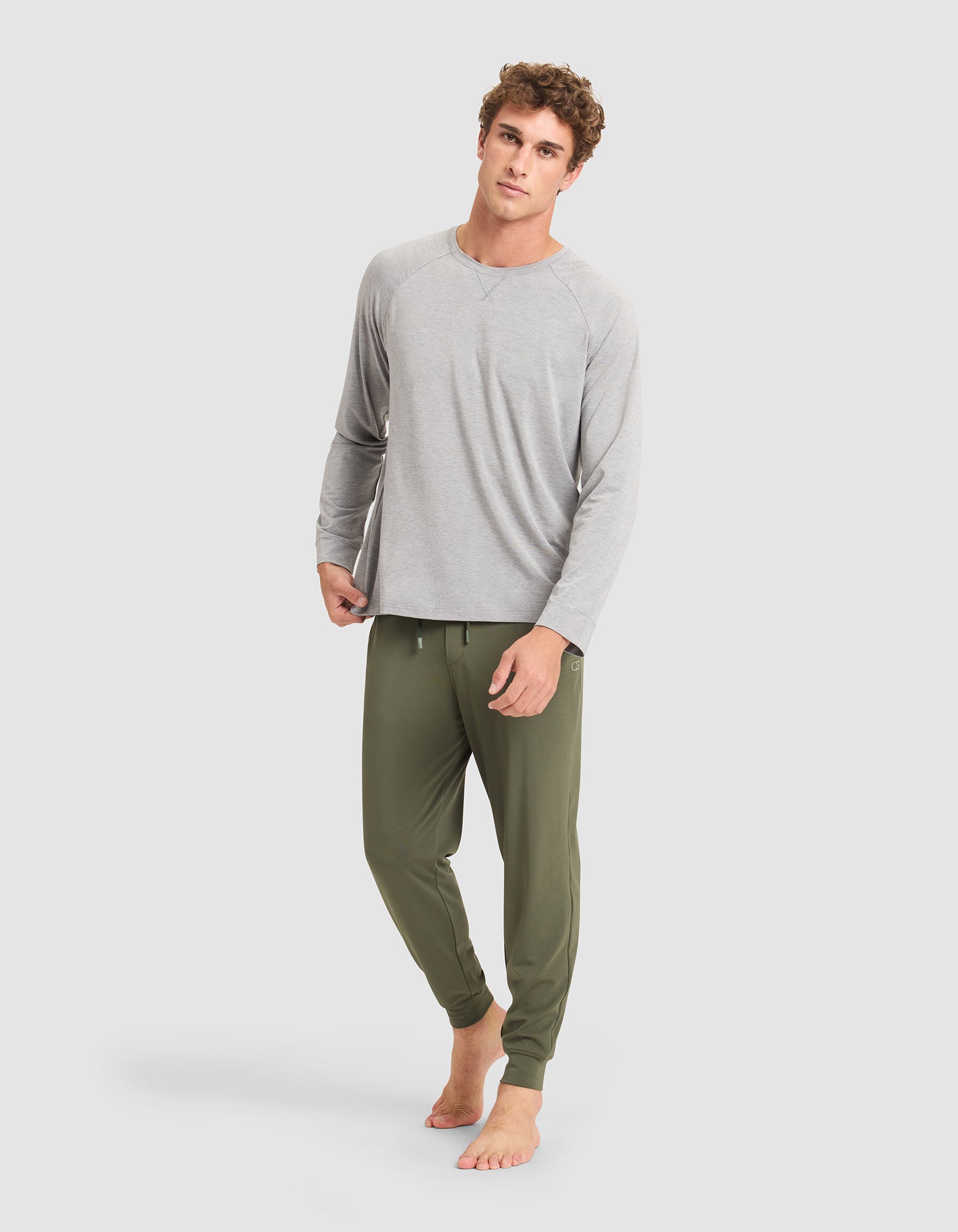 A man stands barefoot against a plain light background wearing the Cozy Earth Men's Bamboo Stretch-Knit Long Sleeve in light gray and olive green jogger pants. 