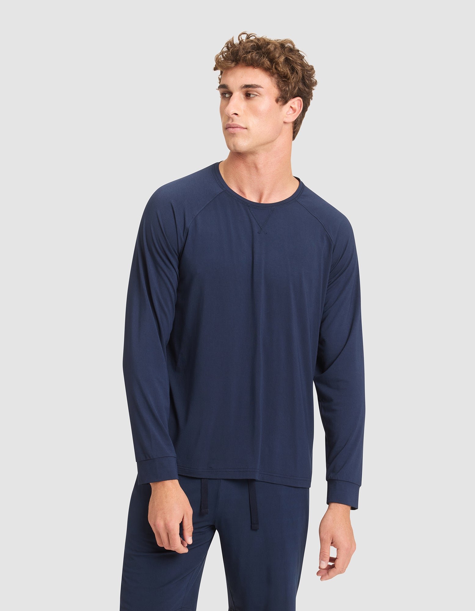 A young man with curly hair wears Cozy Earth's Men's Bamboo Stretch-Knit Long Sleeve in navy blue, paired with matching pants, standing against a plain light gray background and looking slightly to his left.  |Color:Navy
