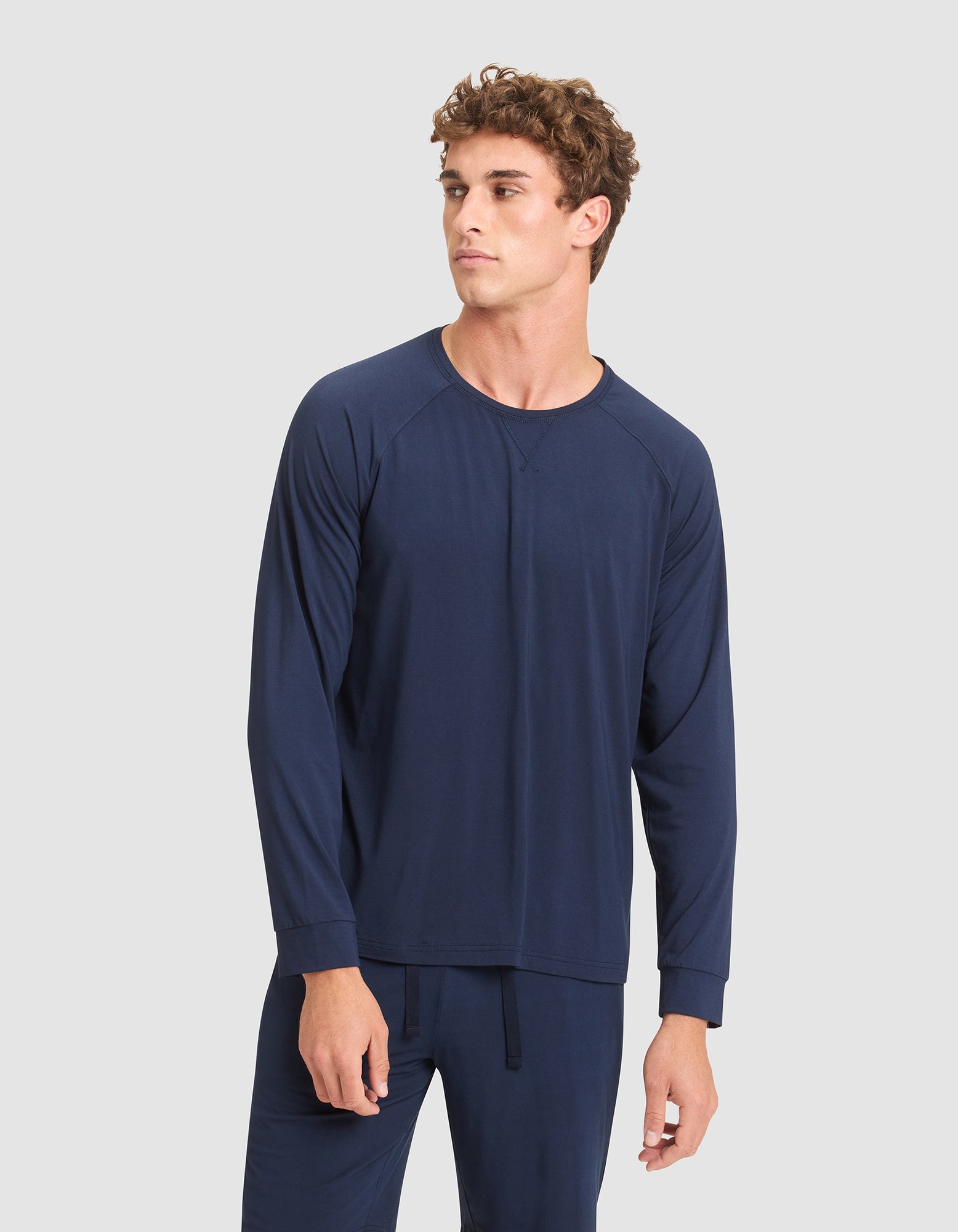 A young man with curly hair wears Cozy Earth's Men's Bamboo Stretch-Knit Long Sleeve in navy blue, paired with matching pants, standing against a plain light gray background and looking slightly to his left.  