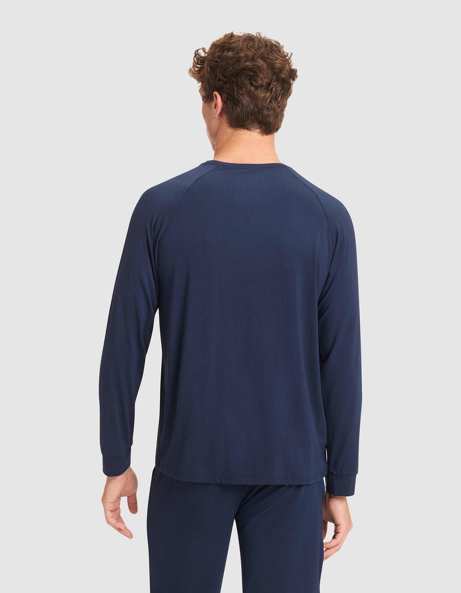 A man with short curly hair faces away from the camera, wearing Cozy Earth's Men's Bamboo Stretch-Knit Long Sleeve in navy blue, paired with matching pants, against a light gray background. |Color:Navy