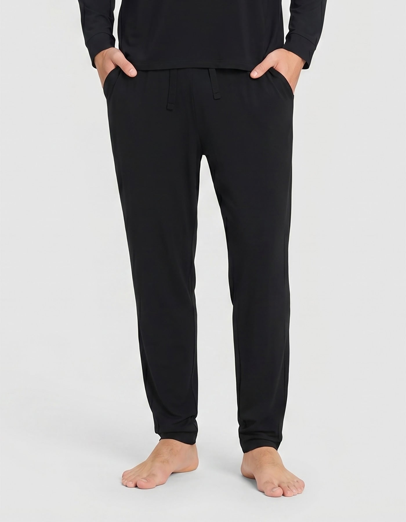 A person stands barefoot against a plain light background, wearing the Cozy Earth Men's Bamboo Stretch-Knit Long Sleeve Pajama Set in black, with hands in their pockets.