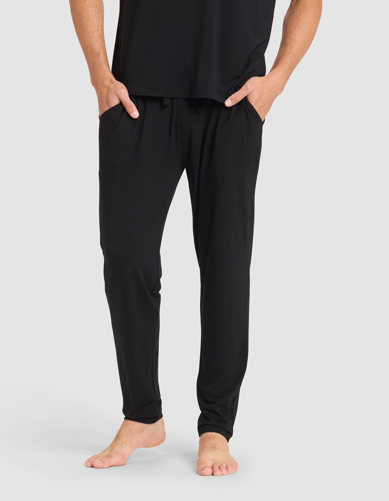 A person stands barefoot with hands in pockets, wearing Cozy Earth's Men's Bamboo Stretch-Knit Pajama Pant in black and a matching black shirt against a plain light gray background. The image is cropped at the shoulders. 