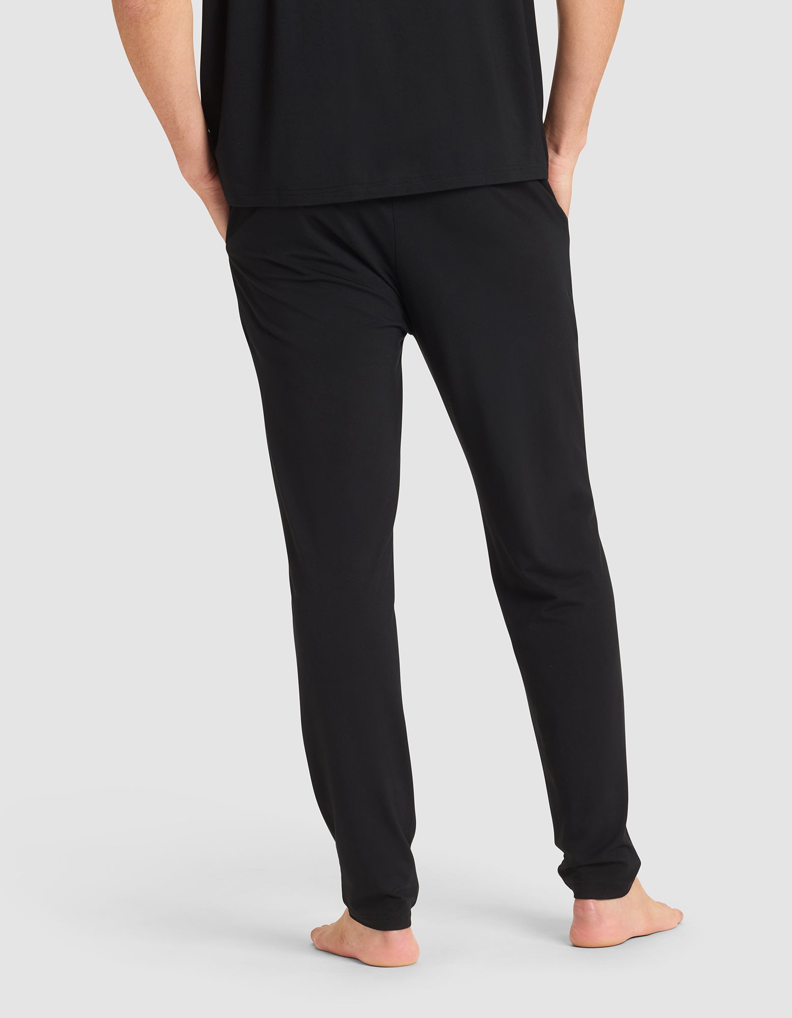 A person shown from the back, barefoot with hands in pockets, wearing Cozy Earth's Men's Bamboo Stretch-Knit Pajama Pant in black and a matching black shirt against a plain light background. 