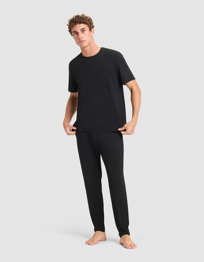 A man stands barefoot against a plain background, wearing Cozy Earth's black Bamboo Stretch-Knit Pajama Pant with a matching black short-sleeve t-shirt featuring a chest pocket.