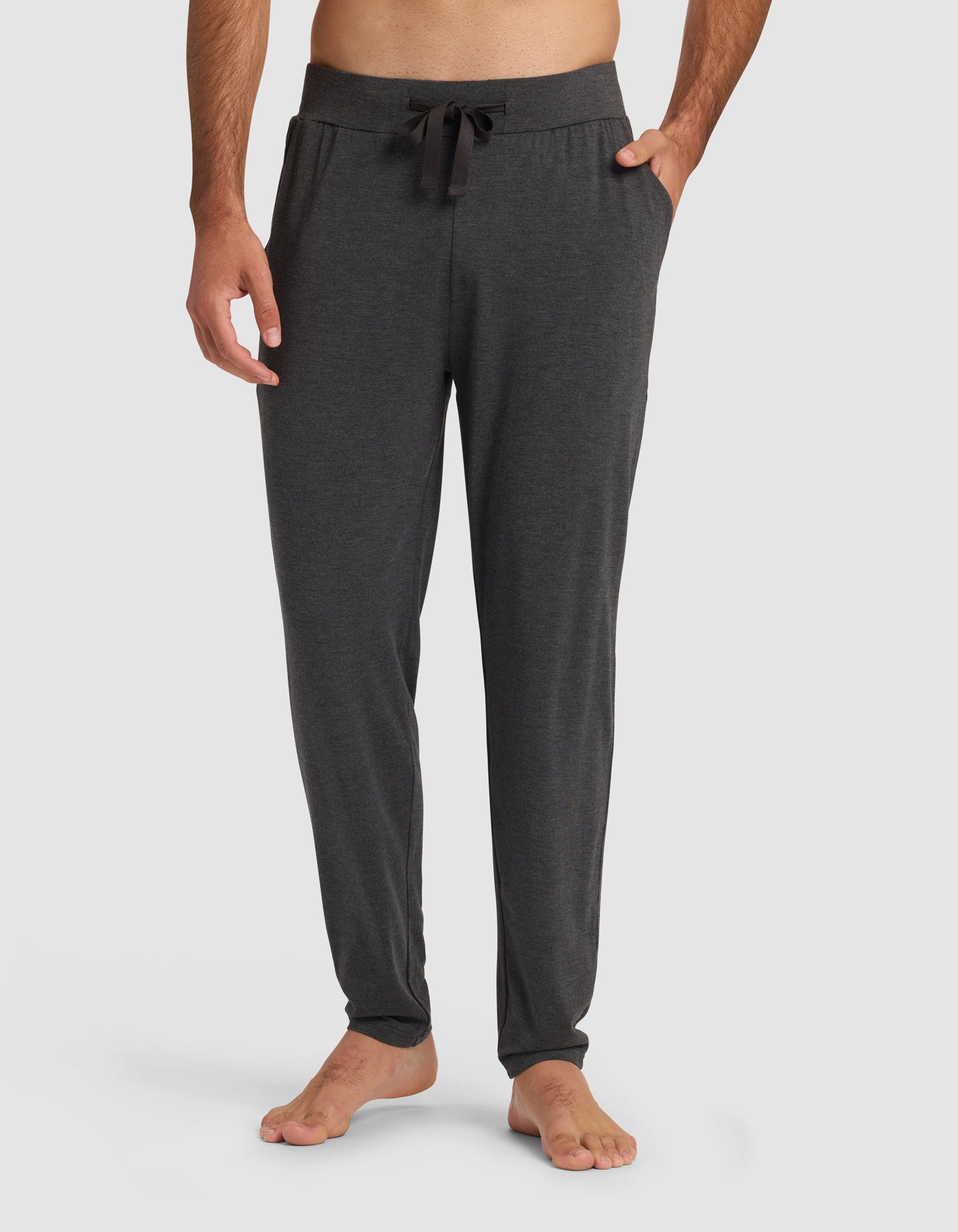 A man stands barefoot with hands in his pockets, wearing Cozy Earth's Men's Bamboo Stretch-Knit Pajama Pant in dark gray, featuring a drawstring waistband, against a plain light background.|Color:Charcoal