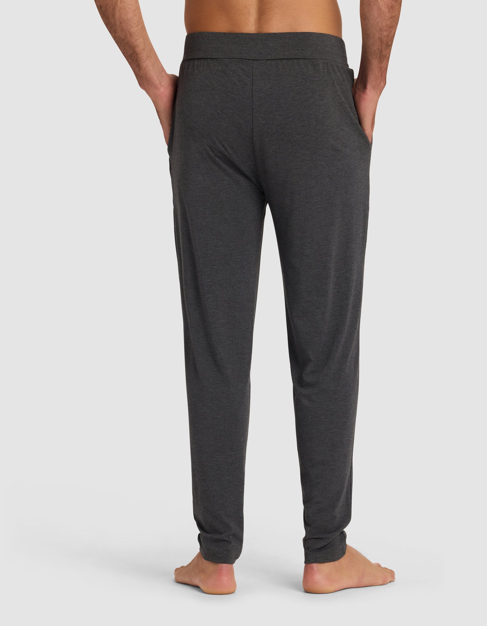A man stands barefoot with his hands in his pockets, wearing Cozy Earth Men's Bamboo Stretch-Knit Pajama Pants in dark gray, viewed from the back against a plain light background.|Color:Charcoal