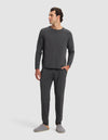 A man with short dark hair smiles in a plain white setting, wearing Cozy Earth Men's Bamboo Stretch-Knit Pajama Pants and matching dark gray long-sleeve top, paired with gray slippers and one hand in his pocket.|Color:Charcoal