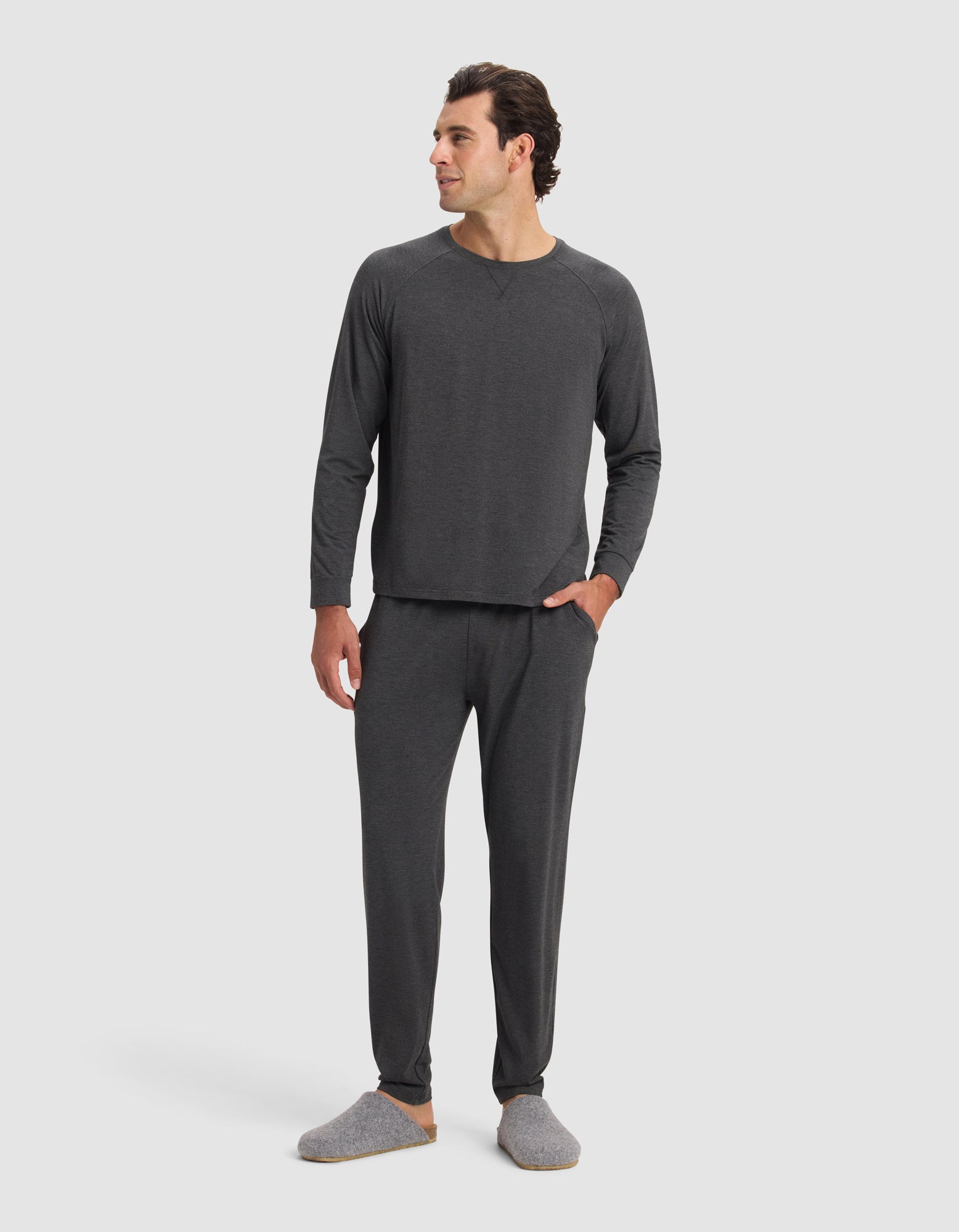 A man with short dark hair smiles in a plain white setting, wearing Cozy Earth Men's Bamboo Stretch-Knit Pajama Pants and matching dark gray long-sleeve top, paired with gray slippers and one hand in his pocket.