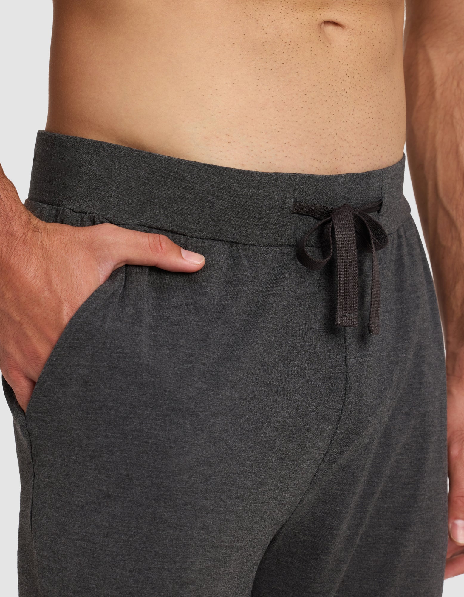 A close-up of a person wearing Cozy Earth Men's Bamboo Stretch-Knit Pajama Pants in dark gray, featuring a drawstring waistband and pockets; their bare torso is visible above the waistline.|Color:Charcoal