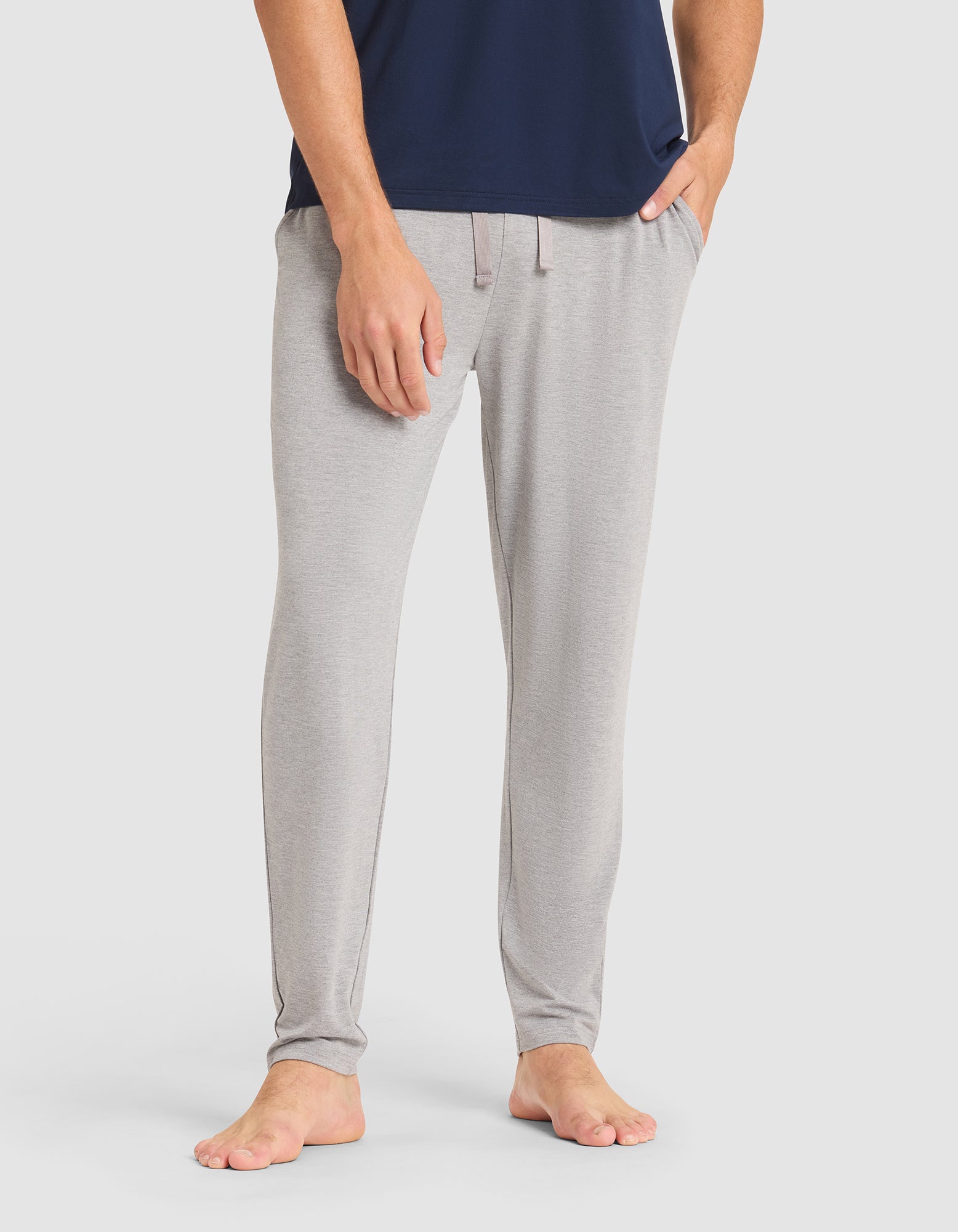 A person is barefoot, wearing Cozy Earth Men's Bamboo Stretch-Knit Pajama Pant in light gray with hands in the pockets, paired with a navy blue shirt. The background is plain white. 