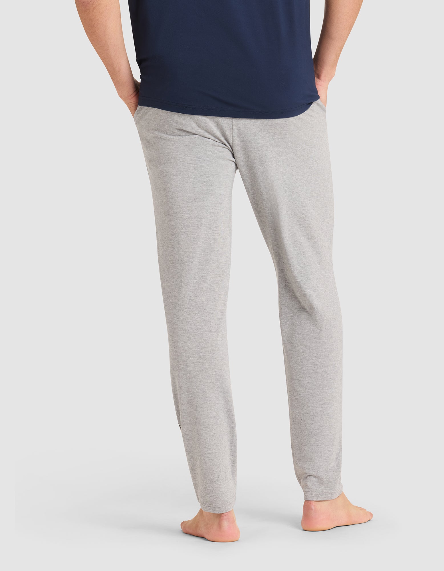 A man stands barefoot with hands in pockets, wearing Cozy Earth's Men's Bamboo Stretch-Knit Pajama Pant in light gray and a dark blue shirt, shown from the back against a plain white background.  