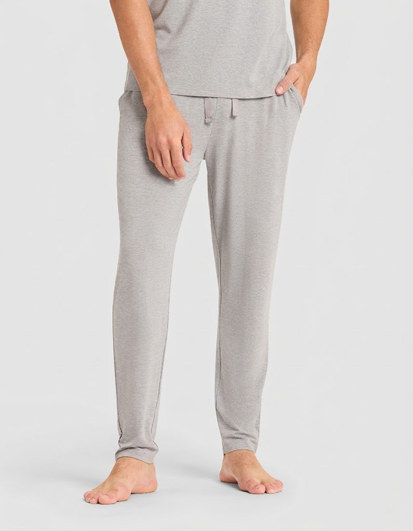 A person models the Cozy Earth Men's Bamboo Stretch-Knit Lounge Tee & Pant Pajama Set in light gray, standing barefoot with one hand in a pocket against a plain light background.