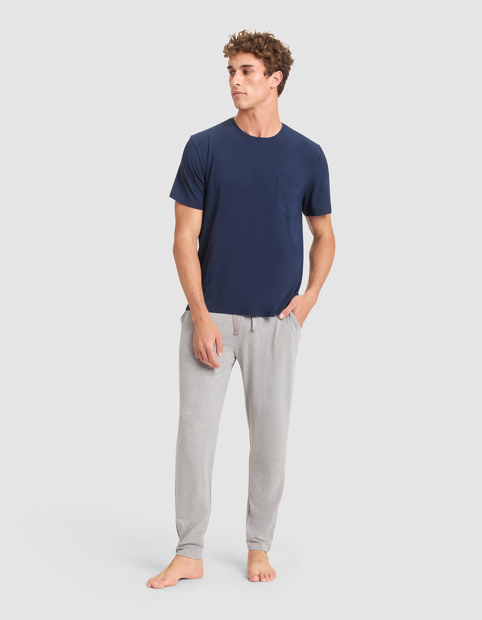 A young man with short curly hair stands barefoot against a plain light gray background, wearing Cozy Earth's Men's Bamboo Stretch-Knit Pajama Pant in light gray and a navy blue short-sleeve t-shirt with a pocket.  