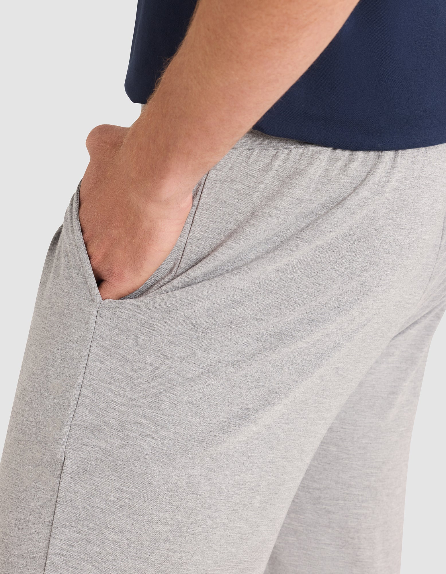 A person is shown from the waist down wearing Cozy Earth Men's Bamboo Stretch-Knit Pajama Pant in light gray with a hand in the pocket, paired with a navy blue shirt, set against a plain background. 