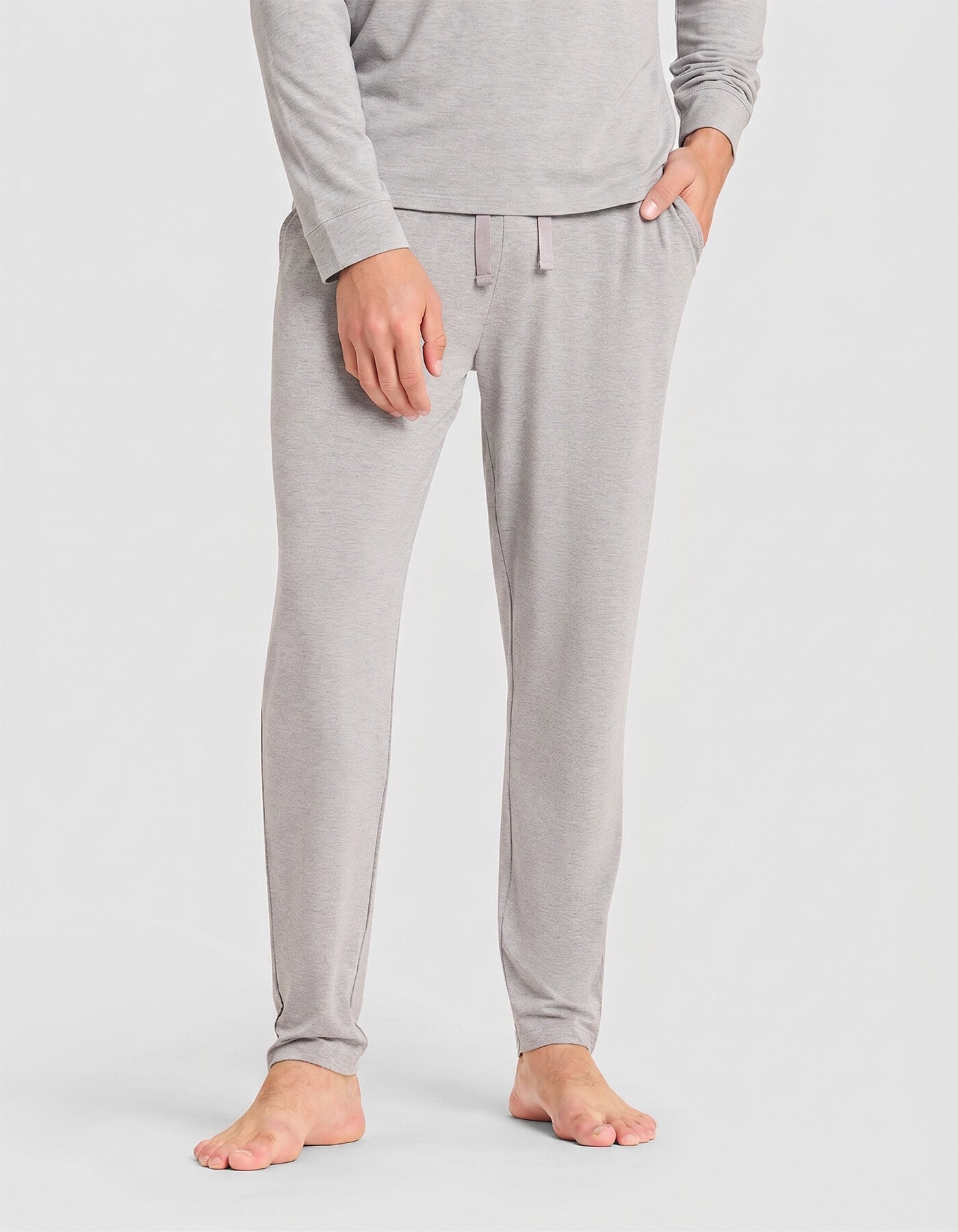 A person stands barefoot, wearing Cozy Earth's Men's Bamboo Stretch-Knit Long Sleeve Pajama Set in light gray; only the lower half of their body is visible against a plain background.