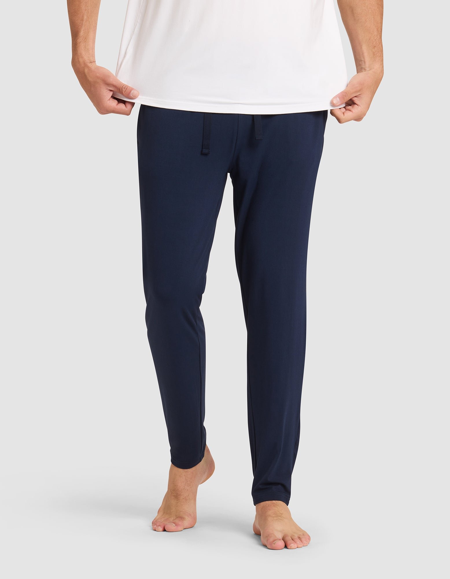 Only the lower half of a person is shown against a plain light background, barefoot and wearing Cozy Earth's Men's Bamboo Stretch-Knit Pajama Pant in navy blue with a drawstring waist and a white shirt, holding the shirt's hem. |Color:Navy
