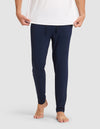 Only the lower half of a person is shown against a plain light background, barefoot and wearing Cozy Earth's Men's Bamboo Stretch-Knit Pajama Pant in navy blue with a drawstring waist and a white shirt, holding the shirt's hem. |Color:Navy