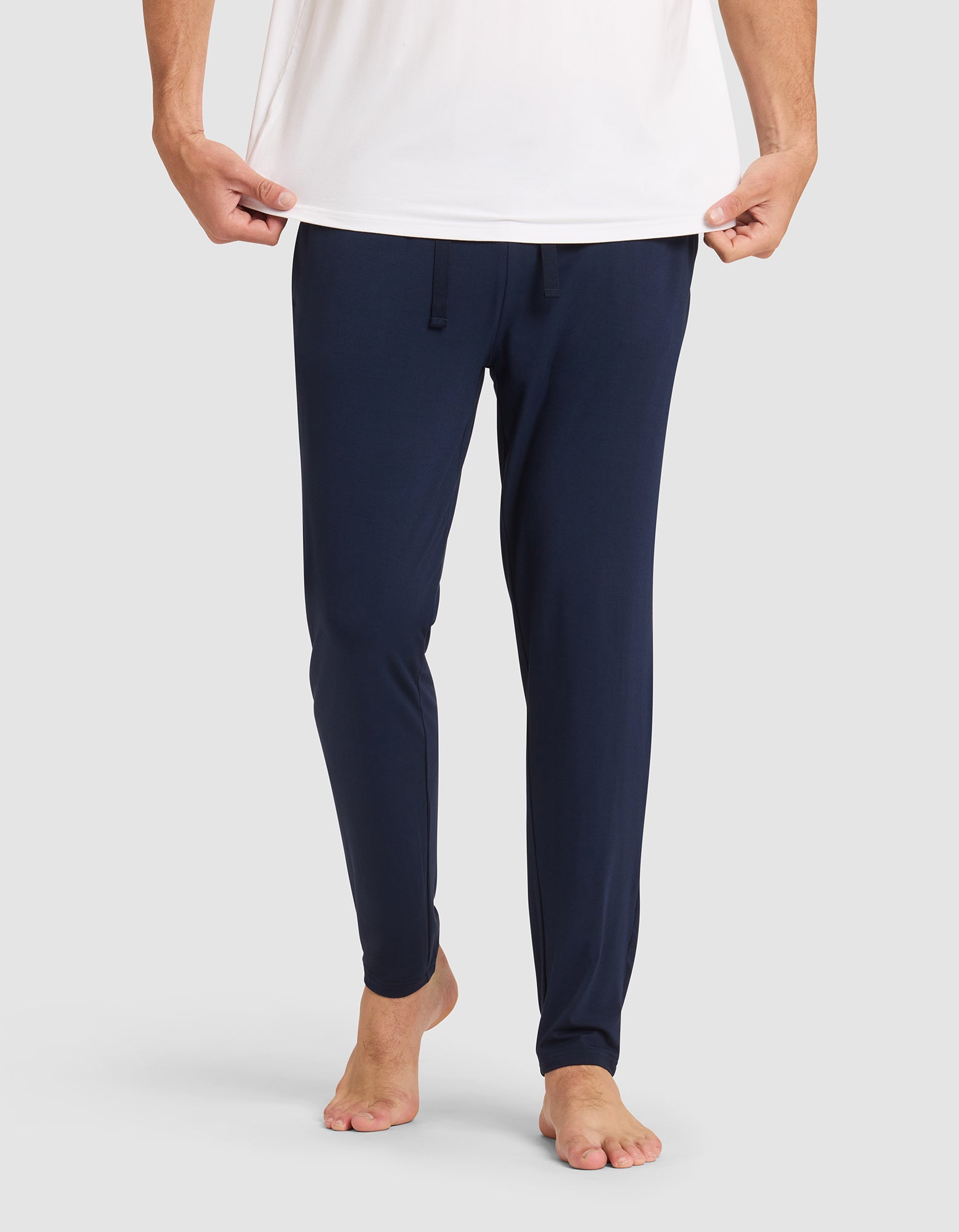 Only the lower half of a person is shown against a plain light background, barefoot and wearing Cozy Earth's Men's Bamboo Stretch-Knit Pajama Pant in navy blue with a drawstring waist and a white shirt, holding the shirt's hem. 