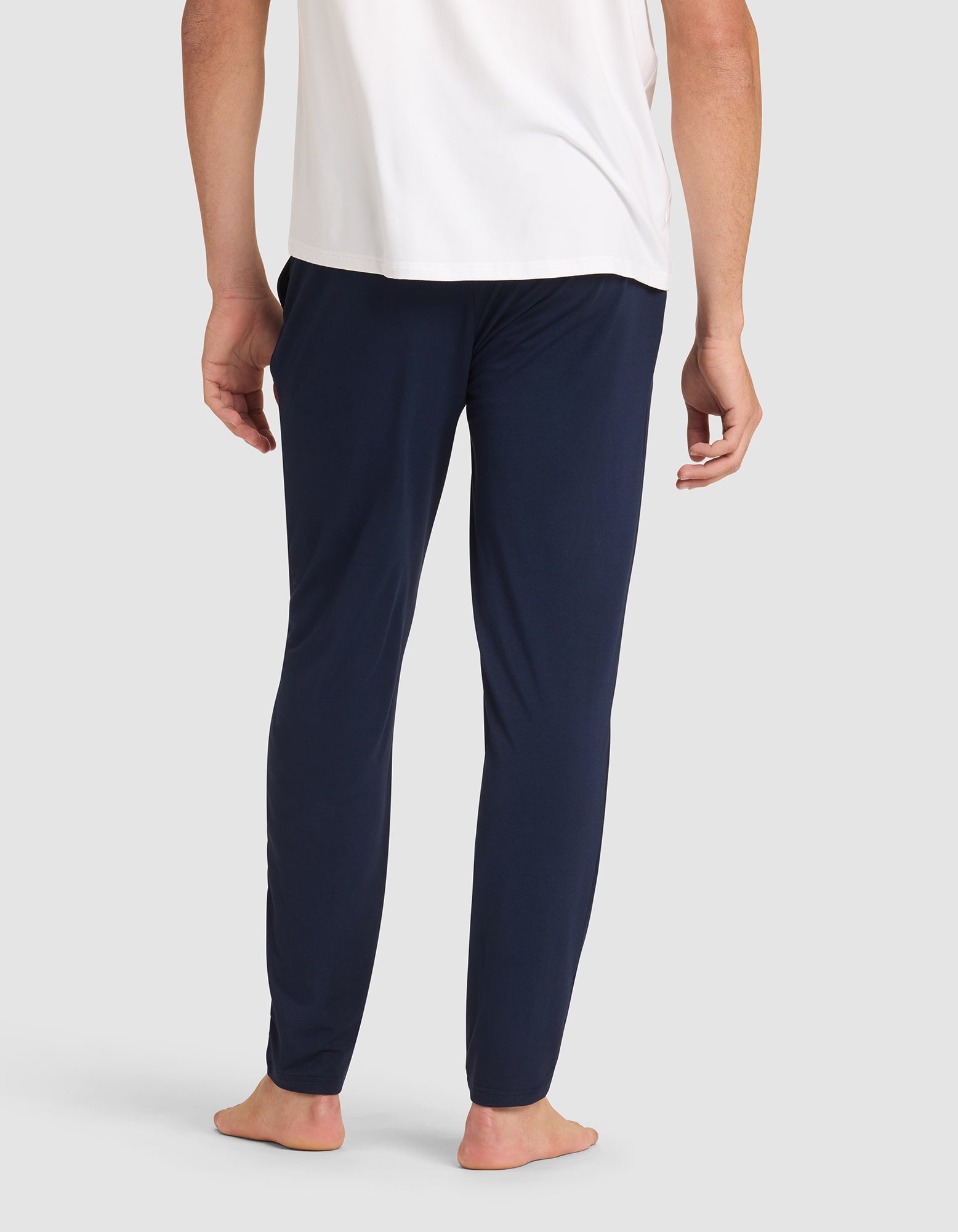 A person stands barefoot, shown from the back, wearing a white shirt and Cozy Earth Men's Bamboo Stretch-Knit Pajama Pant in dark blue against a plain light background. 