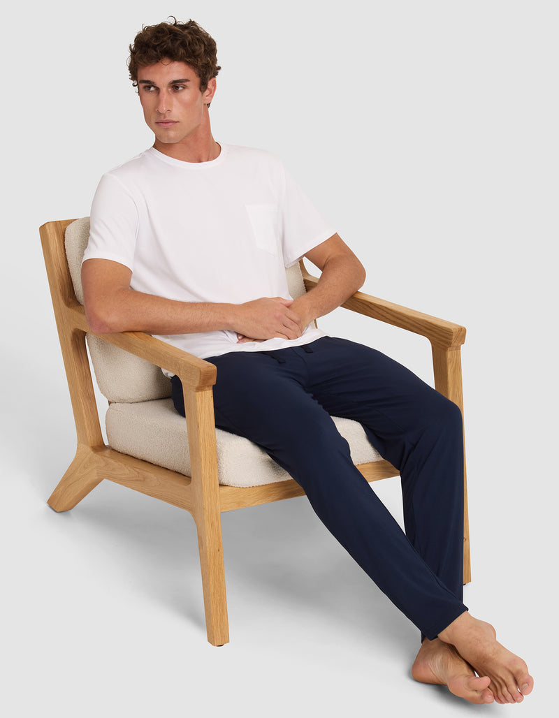 A man with short curly hair relaxes barefoot on a cushioned wooden chair, wearing a white T-shirt and Cozy Earth's Men's Bamboo Stretch-Knit Pajama Pant in navy blue, looking to his left with an easygoing expression.