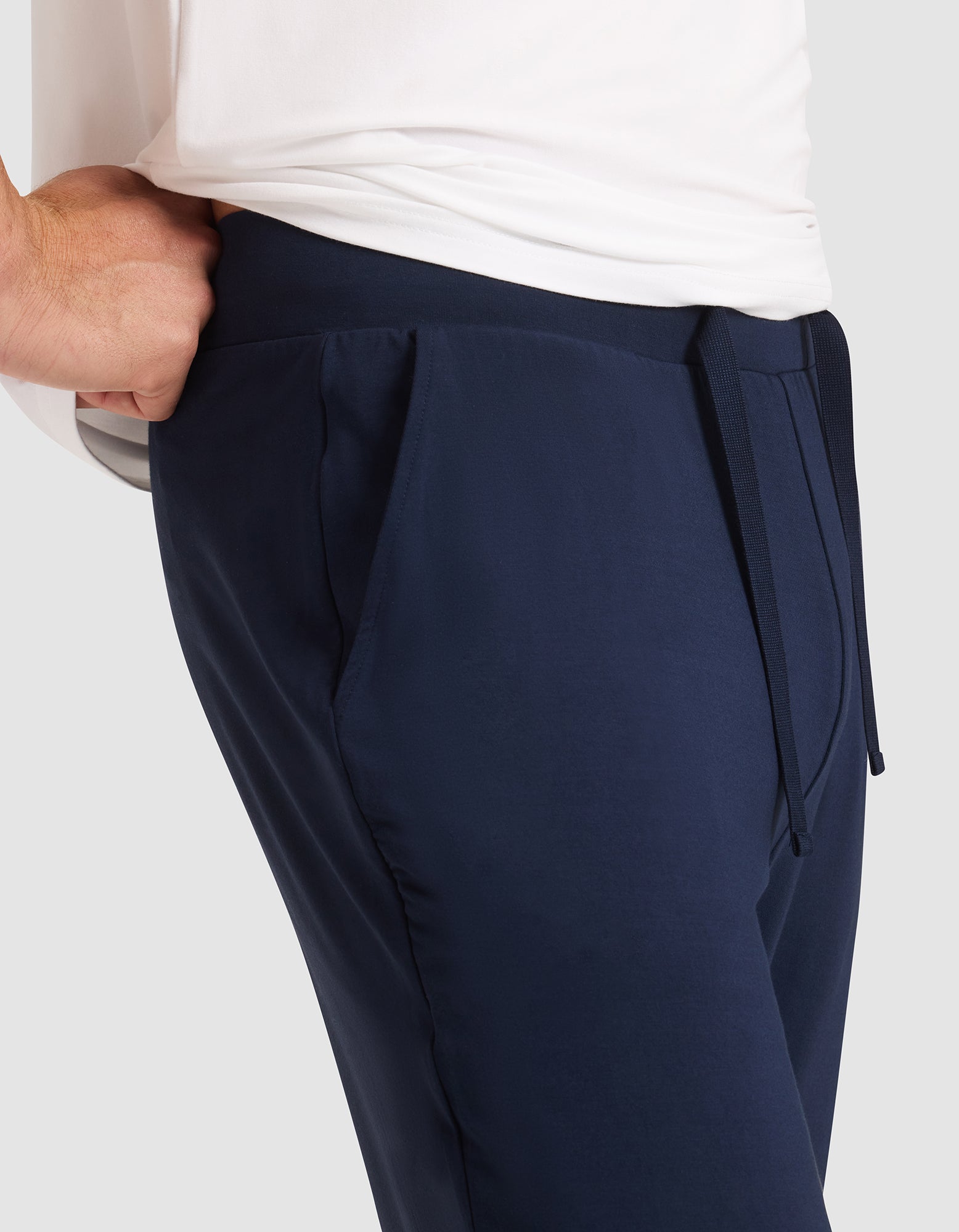 A man wearing Cozy Earth's Men's Bamboo Stretch-Knit Pajama Pant in navy, shown from the side with one hand in his pocket and a visible waist drawstring, paired with a white shirt. 
