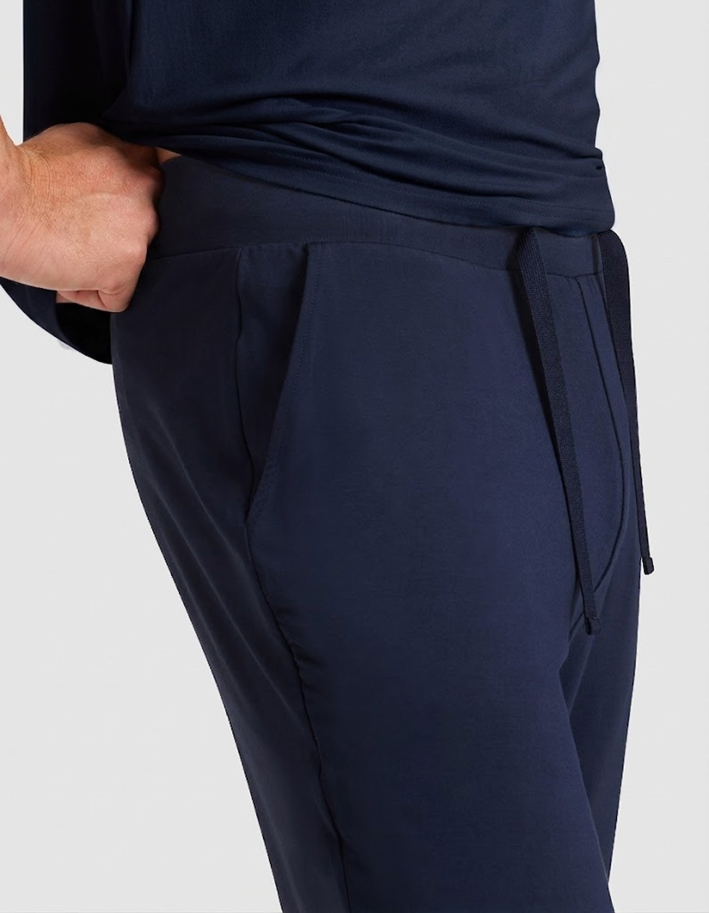 A person wearing Cozy Earth Men's Bamboo Stretch-Knit Lounge Tee & Pant Pajama Set, featuring dark blue pants with a drawstring waistband and their right hand in the pocket, shown from the waist down against a plain background.