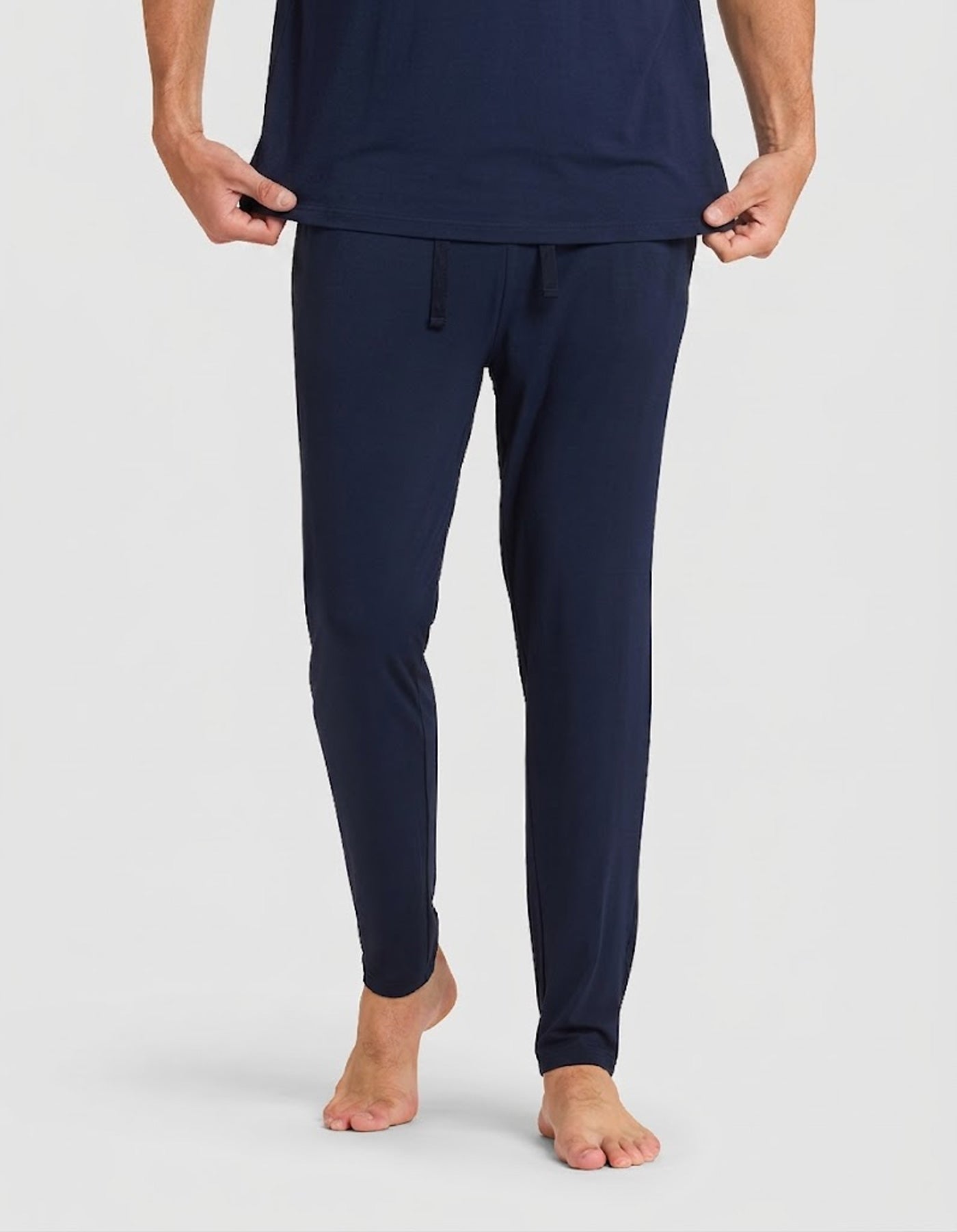 A person stands barefoot in a Cozy Earth Men's Bamboo Stretch-Knit Lounge Tee & Pant Pajama Set in navy blue, holding the shirt slightly lifted against a plain white background.