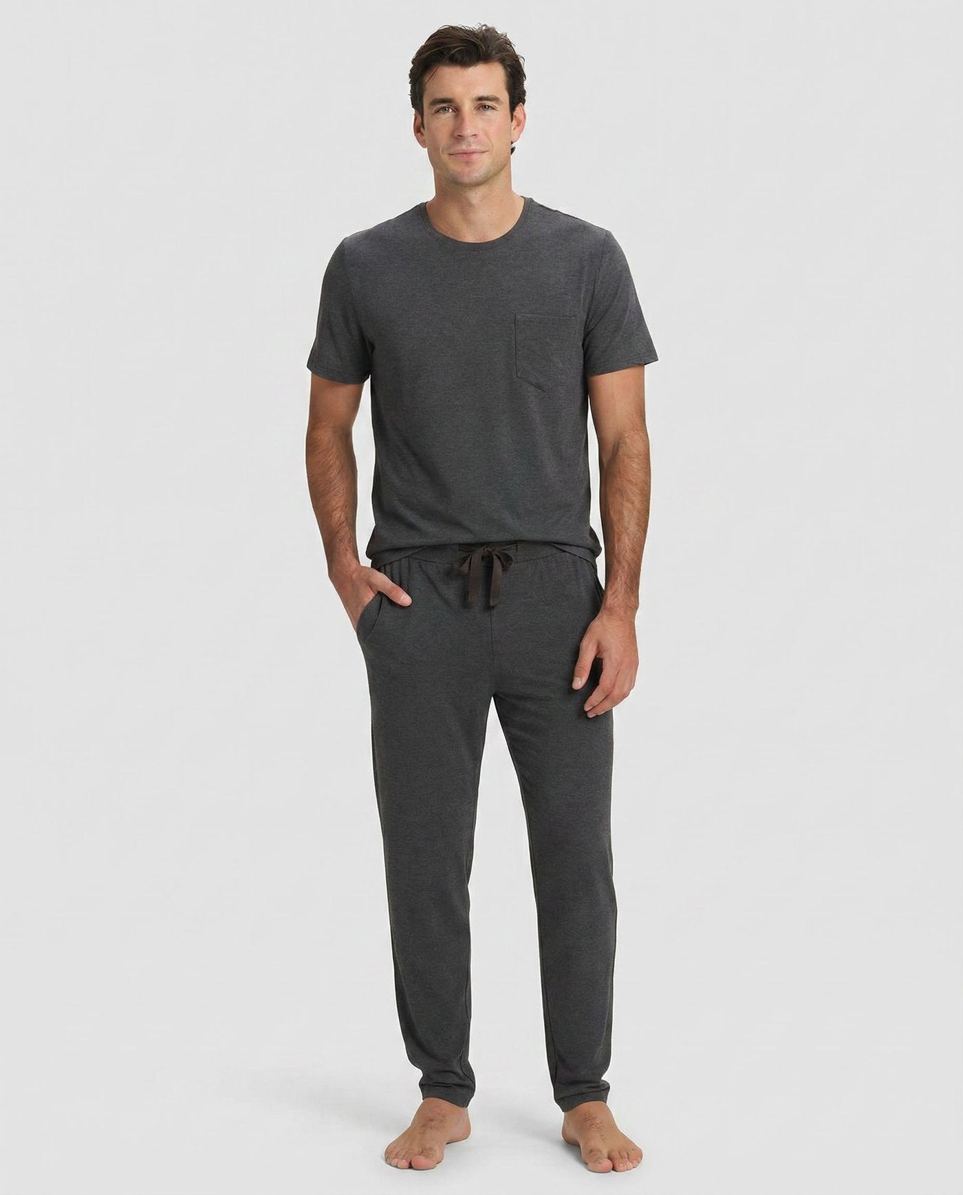 A man stands barefoot against a plain light background, wearing Cozy Earth’s Men's Bamboo Stretch-Knit Lounge Tee & Pant Pajama Set in Charcoal.