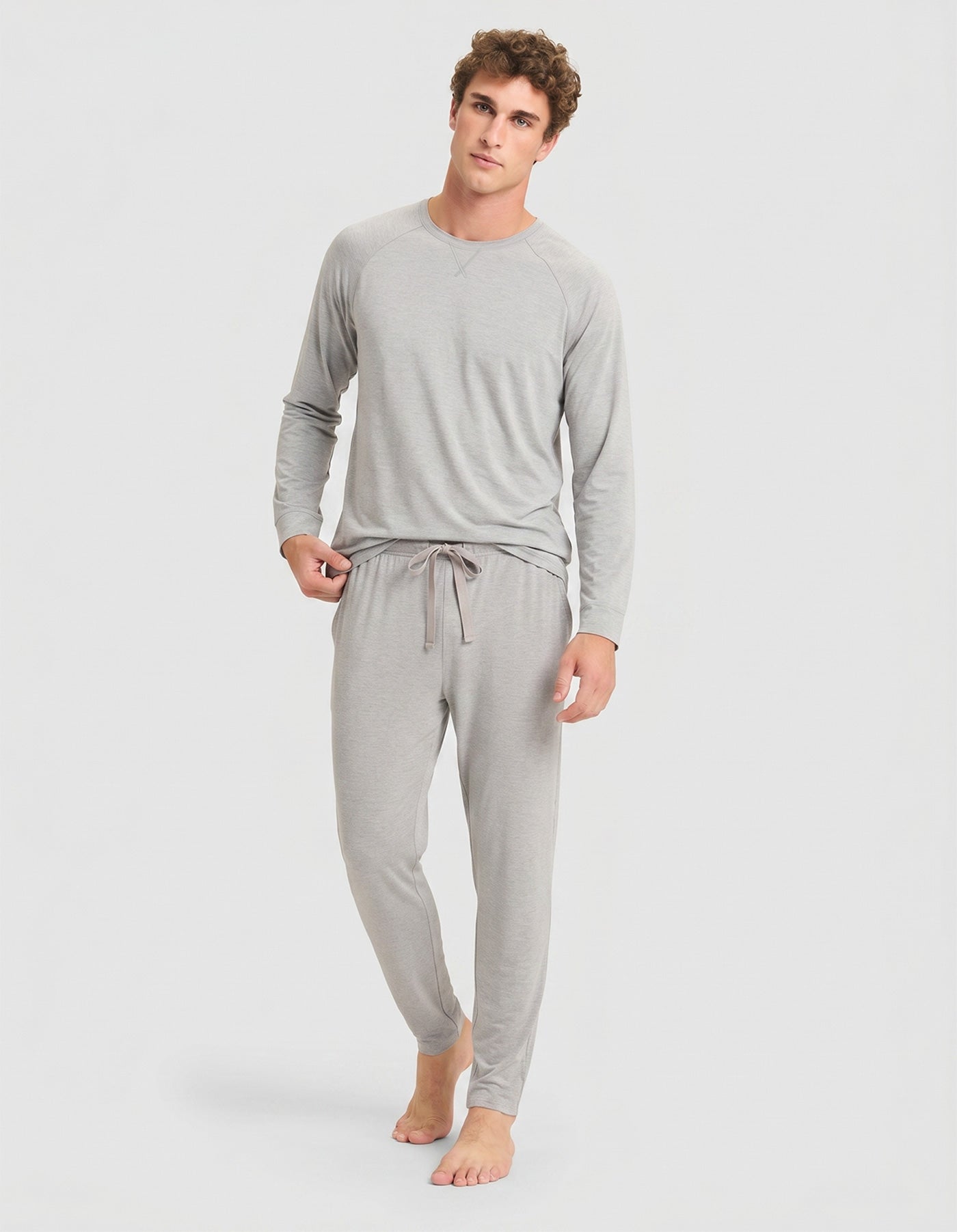 A person stands barefoot against a plain light background, wearing the Cozy Earth Men's Bamboo Stretch-Knit Long Sleeve Pajama Set in Heather Grey.