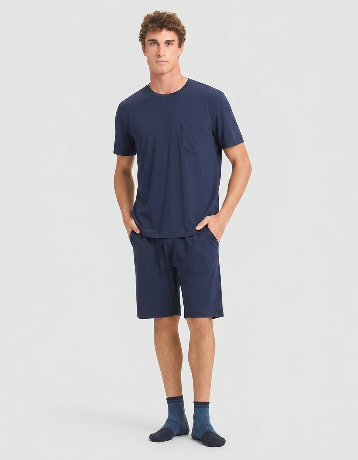 A person stands against a plain background wearing the Cozy Earth Men's Bamboo Stretch-Knit Short Sleeve Pajama Set in navy, with hands in the shorts' pockets.