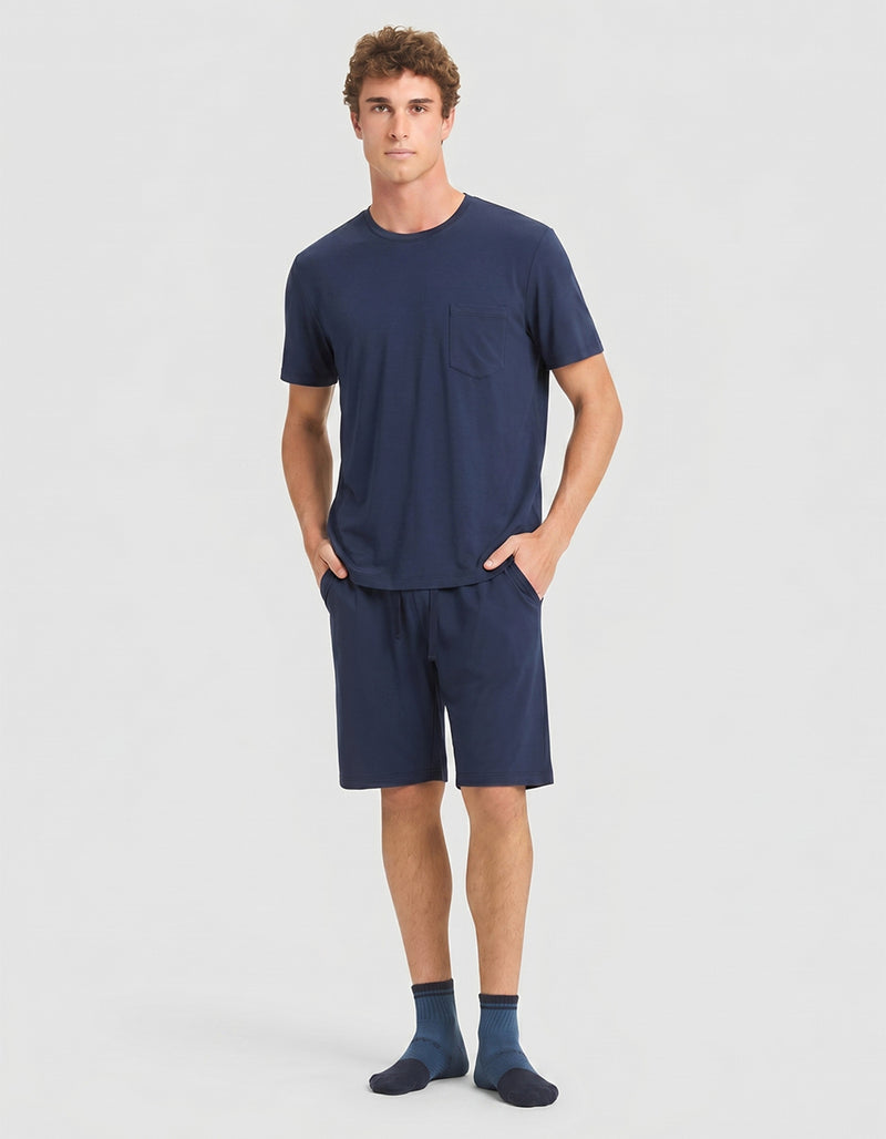 A person stands against a plain background wearing the Cozy Earth Men's Bamboo Stretch-Knit Short Sleeve Pajama Set in navy, with hands in the shorts' pockets.