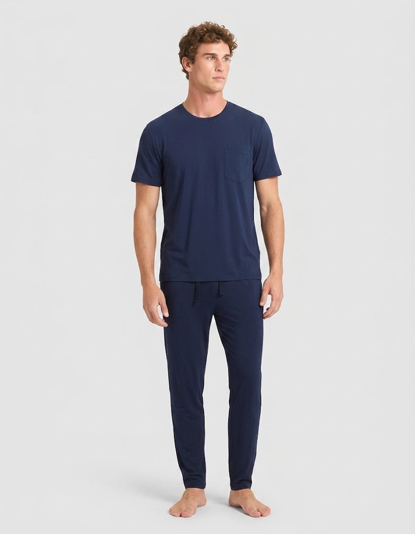 A man stands barefoot against a light gray background, wearing Cozy Earth’s Men's Bamboo Stretch-Knit Lounge Tee & Pant Pajama Set in navy.