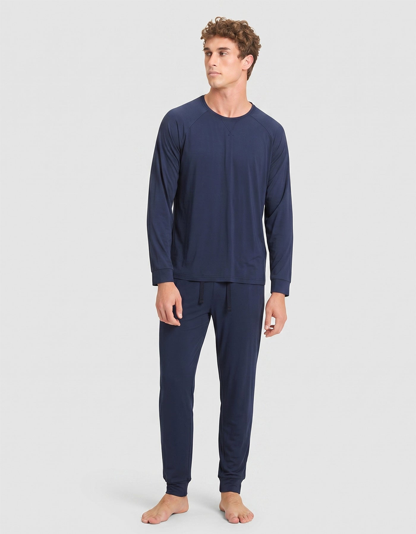 A young man with curly hair wears Cozy Earth's Men's Bamboo Stretch-Knit Long Sleeve Pajama Set in navy blue, standing against a plain light gray background and looking slightly to his left.  